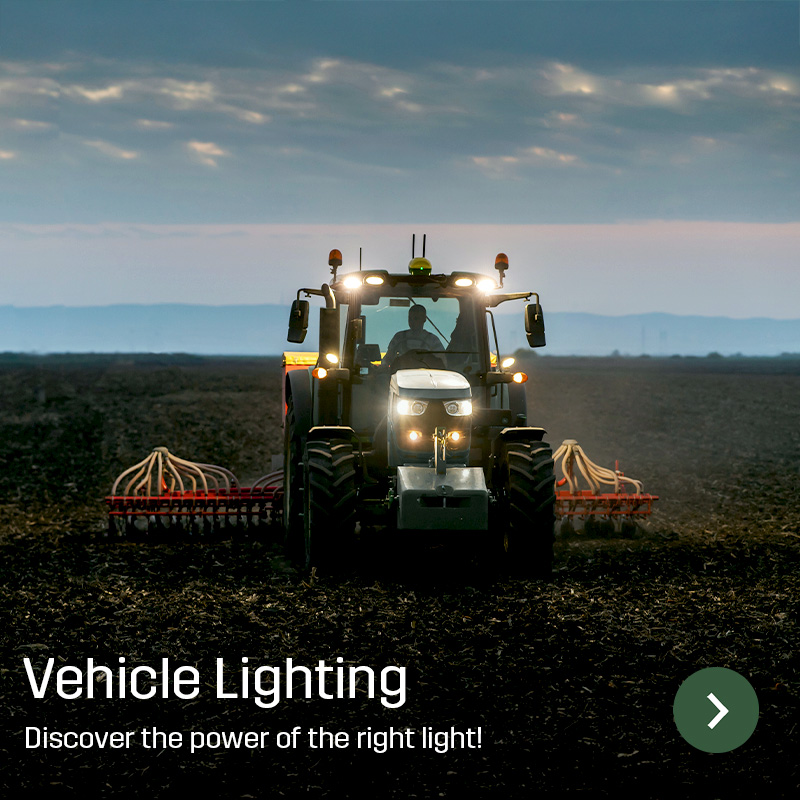 Tractor with work lights switched on driving across farmland with a wide implement behind. Sagros vehicle lighting provides illumination around agricultural machinery during field work.