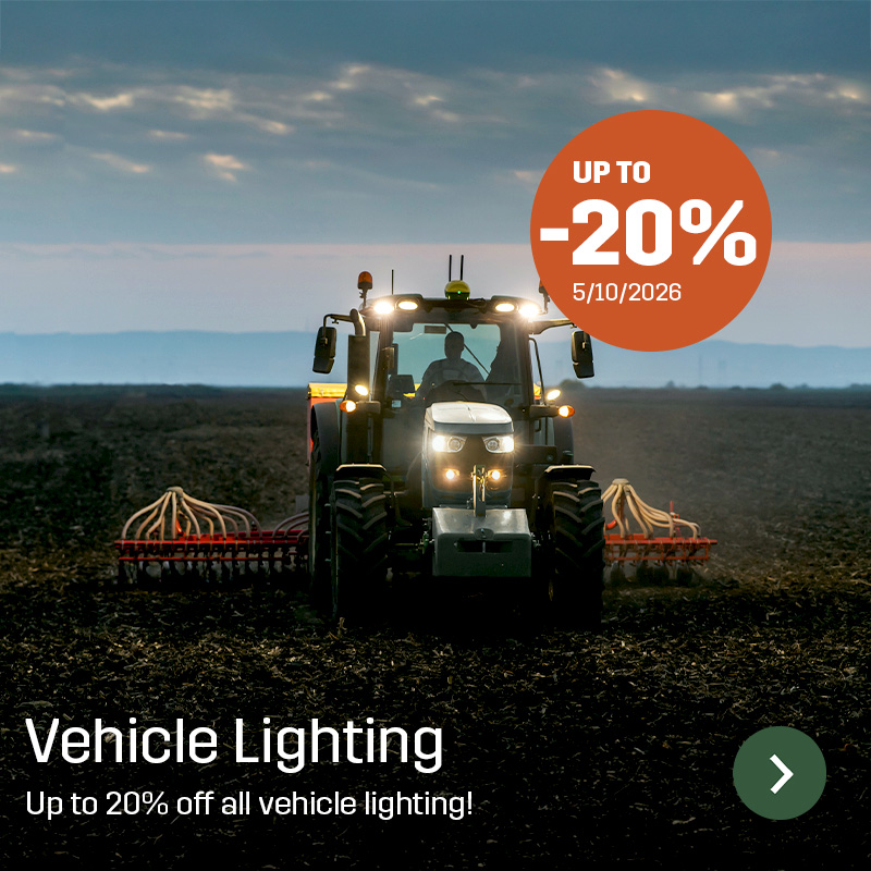 Tractor with work lights switched on driving across farmland with a wide implement behind. Sagros vehicle lighting provides illumination around agricultural machinery during field work.