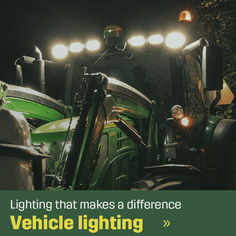 Close-up of a green tractor with a front loader, illuminated LED work lights on the cab roof and amber warning beacons in the dark. Sagros vehicle lighting range includes work lights and warning beacons for tractors and agricultural machinery.